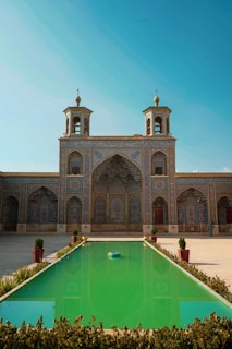 A majestic Persian-style architectural building with intricate tile work in blue and gold hues. The structure features two symmetrical minarets and an expansive arched facade. In the forefront, a long rectangular pool with a vibrant green tint reflects the building. The pool is flanked by planters containing small shrubs, enhancing the tranquil and grand ambiance.