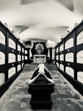 A black and white image depicting an interior with a central black coffin adorned with white fringes. The walls are lined with niches and plaques, possibly identifying remembered individuals. At the far end of the space, there is an ornate altar or religious display with intricate detailing.