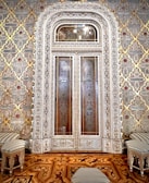 An intricately designed door with ornate carvings and patterns dominates the center of the image. The door is flanked by an elaborate wallpaper with geometric and floral motifs in gold, red, and beige. In front of the door, there are striped upholstered stools and a small table with similar design elements. The floor features a rich wooden parquet with an intricate inlay design.