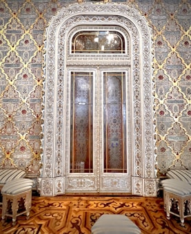 An intricately designed door with ornate carvings and patterns dominates the center of the image. The door is flanked by an elaborate wallpaper with geometric and floral motifs in gold, red, and beige. In front of the door, there are striped upholstered stools and a small table with similar design elements. The floor features a rich wooden parquet with an intricate inlay design.