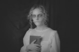 A person with a ghostly appearance is holding a book. They have pale skin, hollow eyes, and dark makeup emphasizing a skeletal look. The image is in black and white, adding to a mysterious and eerie atmosphere.