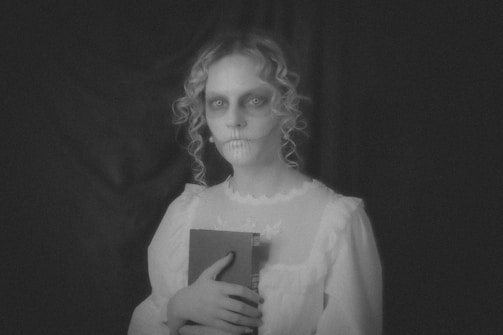 A person with a ghostly appearance is holding a book. They have pale skin, hollow eyes, and dark makeup emphasizing a skeletal look. The image is in black and white, adding to a mysterious and eerie atmosphere.