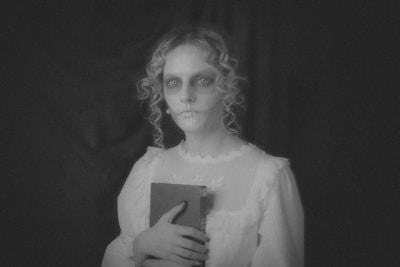 A person with a ghostly appearance is holding a book. They have pale skin, hollow eyes, and dark makeup emphasizing a skeletal look. The image is in black and white, adding to a mysterious and eerie atmosphere.