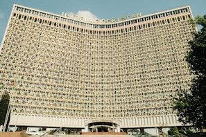 A large hotel building with a concave facade covered in numerous identical rectangular windows. The structure is prominently labeled 'Hotel Uzbekistan' at the top and has a symmetrical, grid-like pattern.