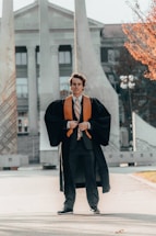 A person wearing graduation attire, featuring a black gown with an orange stole, stands on a pathway in front of a monumental structure with columns. The background shows a large building with columns and windows, set in a sunny area with some trees visible.