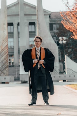 A person wearing graduation attire, featuring a black gown with an orange stole, stands on a pathway in front of a monumental structure with columns. The background shows a large building with columns and windows, set in a sunny area with some trees visible.