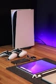 A sleek, modern setup featuring a gaming console with two controllers on a wooden surface. Nearby are a smartphone, smartwatch, tablet with a vivid screen, wireless earbuds, and a stylus. The background is illuminated with a soft purple glow.