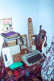 A vintage typewriter is placed on a colorful patterned tablecloth. Nearby, there are various books, one of which is titled 'Emotional Intelligence'. There is also a bottle filled with white capsules or small items, and an abstract piece of artwork. Decorative objects, including a wooden board game and animal figurines, are part of the setup, giving a cozy and eclectic vibe.
