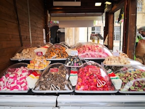 A market stall displaying a variety of colorful sweets is set against a wooden backdrop. Plastic trays hold cubes of fudge in various flavors and colors, covered with clear plastic domes. Small signs label each type, such as caramel, licorice, and assorted fruit flavors. A person stands off to the side, partially visible wearing a hat and jacket.