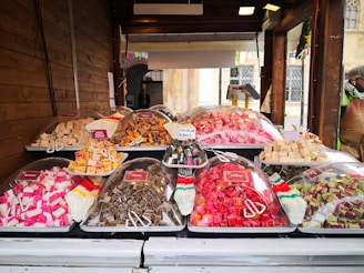 A market stall displaying a variety of colorful sweets is set against a wooden backdrop. Plastic trays hold cubes of fudge in various flavors and colors, covered with clear plastic domes. Small signs label each type, such as caramel, licorice, and assorted fruit flavors. A person stands off to the side, partially visible wearing a hat and jacket.