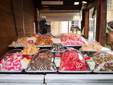 A market stall displaying a variety of colorful sweets is set against a wooden backdrop. Plastic trays hold cubes of fudge in various flavors and colors, covered with clear plastic domes. Small signs label each type, such as caramel, licorice, and assorted fruit flavors. A person stands off to the side, partially visible wearing a hat and jacket.