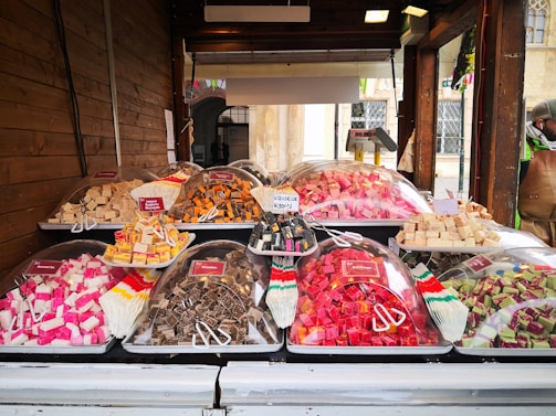 A market stall displaying a variety of colorful sweets is set against a wooden backdrop. Plastic trays hold cubes of fudge in various flavors and colors, covered with clear plastic domes. Small signs label each type, such as caramel, licorice, and assorted fruit flavors. A person stands off to the side, partially visible wearing a hat and jacket.