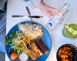 A plate filled with grilled fish, rice, and a fresh salad composed of greens, tomatoes, and spiralized vegetables. A small container of dressing is on the side. Next to the plate, there is a bottle of sparkling water being held by a hand with blue-painted nails. A drink with cucumber slices arranged on top sits nearby, accompanied by a bowl of seasoned shrimp.