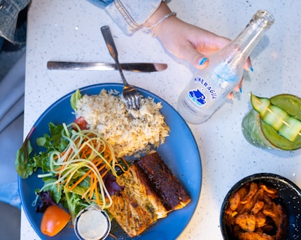 A plate filled with grilled fish, rice, and a fresh salad composed of greens, tomatoes, and spiralized vegetables. A small container of dressing is on the side. Next to the plate, there is a bottle of sparkling water being held by a hand with blue-painted nails. A drink with cucumber slices arranged on top sits nearby, accompanied by a bowl of seasoned shrimp.