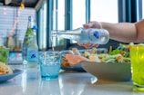 A hand pours sparkling water from a bottle into a blue glass placed on a table. The table is set with a fresh salad, lime drink, and other food dishes under bright natural light, with large windows in the background.