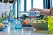 A hand pours sparkling water from a bottle into a blue glass placed on a table. The table is set with a fresh salad, lime drink, and other food dishes under bright natural light, with large windows in the background.
