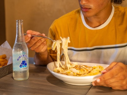 A person wearing a yellow and white striped shirt is sitting at a table with a plate of pasta topped with grilled chicken and Alfredo sauce. The person is holding a fork twirling the pasta. On the table to the left, there is a bottle of Mineragua sparkling water. The setting appears to be a casual dining environment.