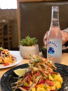A plate of colorful food featuring diced mango, tomatoes, and shredded vegetables sits on a table next to a wedge of lime. In the background, there is a dish topped with what looks like coleslaw and a cheese sauce. A decorative succulent plant is placed in a small pot. A hand is holding a bottle of sparkling water labeled 'Mineragua'. The setting suggests a casual dining atmosphere with wooden furniture visible in the surrounding.