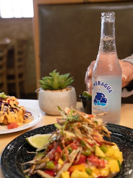 A plate of colorful food featuring diced mango, tomatoes, and shredded vegetables sits on a table next to a wedge of lime. In the background, there is a dish topped with what looks like coleslaw and a cheese sauce. A decorative succulent plant is placed in a small pot. A hand is holding a bottle of sparkling water labeled 'Mineragua'. The setting suggests a casual dining atmosphere with wooden furniture visible in the surrounding.