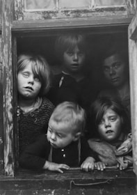 Five children are gathered at a window, looking outward with curious and solemn expressions. The scene is black and white, capturing the texture of worn wood framing the window. The children are dressed in simple, modest clothing.
