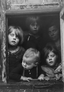 Five children are gathered at a window, looking outward with curious and solemn expressions. The scene is black and white, capturing the texture of worn wood framing the window. The children are dressed in simple, modest clothing.