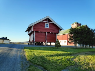 A traditional red wooden barn supported on stilts is situated in a rural area. The barn is surrounded by a well-maintained lawn and a few trees, with another building visible in the background. The scene is bathed in clear daylight under a cloudless blue sky.