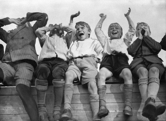 A group of boys sitting on a wooden railing, all appearing joyful and shouting. They are dressed in vintage clothing, including knee socks and shorts. The boys have their arms raised or their hands cupped around their mouths, expressing excitement or enthusiasm.
