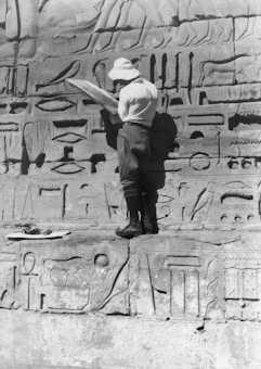 A person wearing a hat and casual clothing stands in front of an ancient stone wall filled with Egyptian hieroglyphs. The individual appears to be studying or recording the hieroglyphs with a large piece of paper or board in hand.