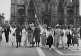 A group of women and men joyfully walking arm in arm, dressed in vintage attire. They are waving flags and carrying large cones, possibly megaphones or cheer horns. The backdrop is a detailed Gothic-style cathedral with ornate architecture.