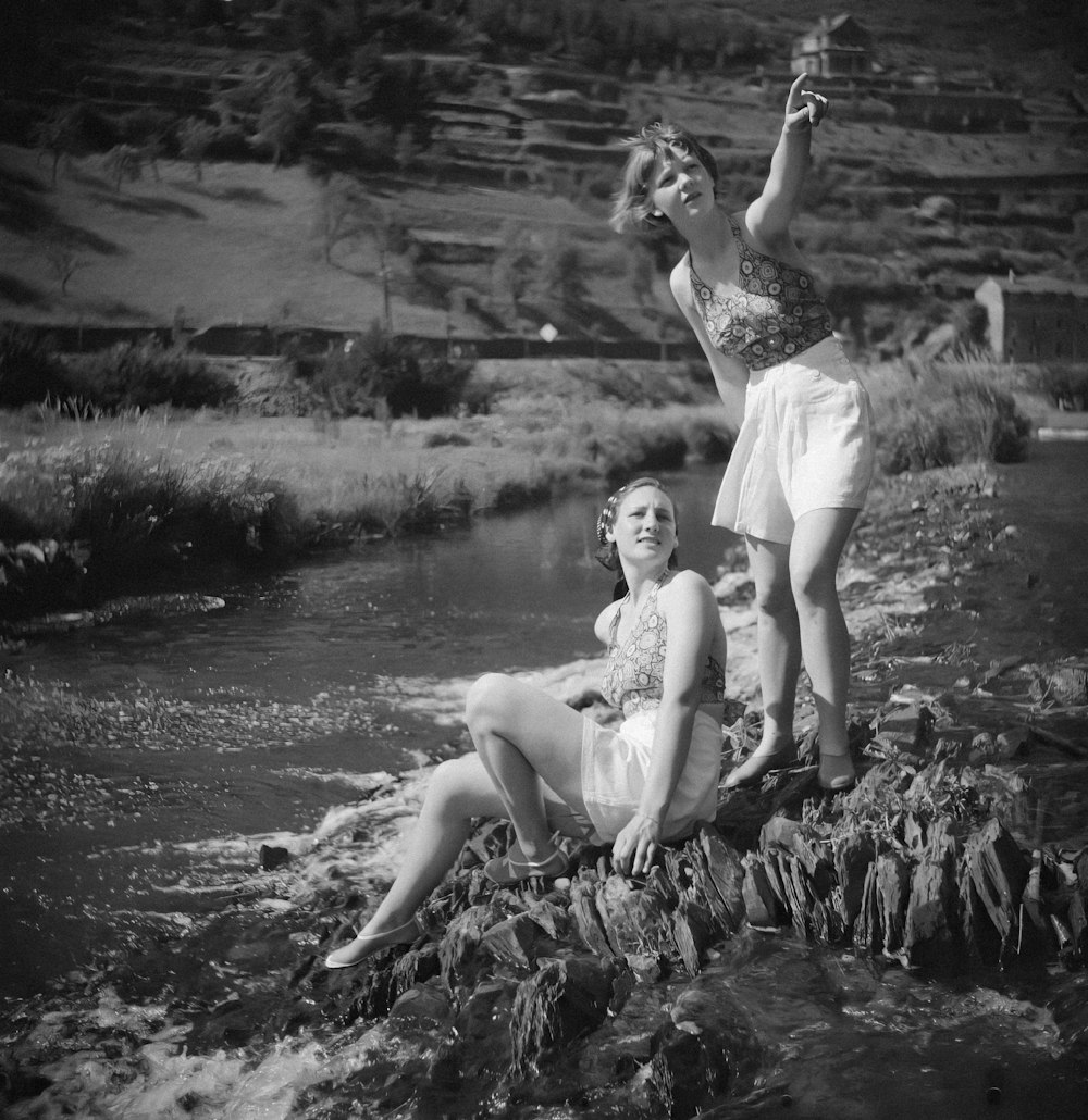 a couple of women sitting on top of a rock next to a river