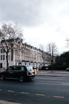 A street scene featuring a row of classic London townhouses with white and beige facades, and a black taxi driving on the road. Leafless trees are visible, indicating a winter or early spring setting. The sky is overcast, casting a muted light over the setting.