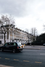 A street scene featuring a row of classic London townhouses with white and beige facades, and a black taxi driving on the road. Leafless trees are visible, indicating a winter or early spring setting. The sky is overcast, casting a muted light over the setting.