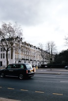 A street scene featuring a row of classic London townhouses with white and beige facades, and a black taxi driving on the road. Leafless trees are visible, indicating a winter or early spring setting. The sky is overcast, casting a muted light over the setting.