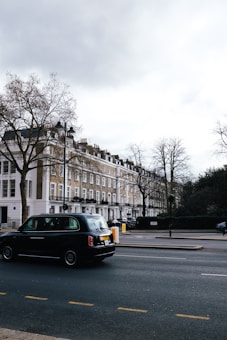 A street scene featuring a row of classic London townhouses with white and beige facades, and a black taxi driving on the road. Leafless trees are visible, indicating a winter or early spring setting. The sky is overcast, casting a muted light over the setting.