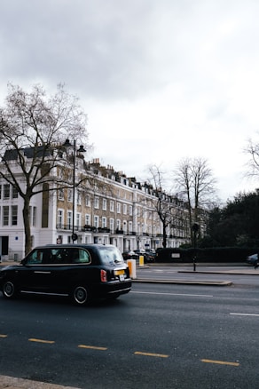 A street scene featuring a row of classic London townhouses with white and beige facades, and a black taxi driving on the road. Leafless trees are visible, indicating a winter or early spring setting. The sky is overcast, casting a muted light over the setting.