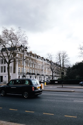 A street scene featuring a row of classic London townhouses with white and beige facades, and a black taxi driving on the road. Leafless trees are visible, indicating a winter or early spring setting. The sky is overcast, casting a muted light over the setting.