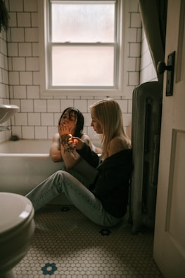 Two people are in a bathroom with white tiled walls and a window above. One person is sitting in a bathtub with wet hair, while the other is sitting on the floor, lighting something. The floor has hexagonal tiles with some blue floral designs.