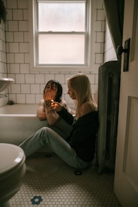 Two people are in a bathroom with white tiled walls and a window above. One person is sitting in a bathtub with wet hair, while the other is sitting on the floor, lighting something. The floor has hexagonal tiles with some blue floral designs.