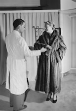 A woman in a luxurious fur coat is conversing with a man in a white coat, possibly a salesperson or tailor, in a room with curtains and a folding screen in the background.