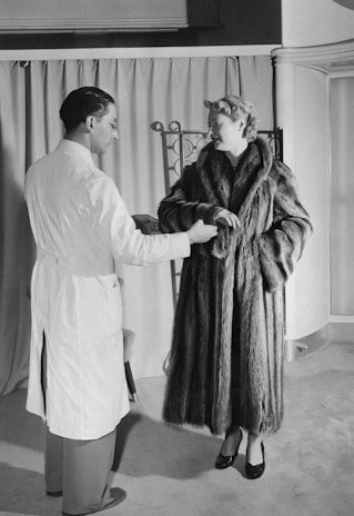 A woman in a luxurious fur coat is conversing with a man in a white coat, possibly a salesperson or tailor, in a room with curtains and a folding screen in the background.