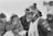 A person dressed for cold weather is covered in snow and ice, wearing a thick knit hat, ski goggles, and a numbered bib. The scene appears to be a winter sports event with spectators visible in the background, bundled in heavy clothing amidst falling snow.