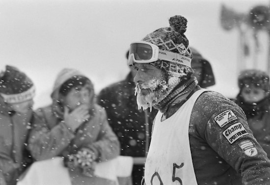 A person dressed for cold weather is covered in snow and ice, wearing a thick knit hat, ski goggles, and a numbered bib. The scene appears to be a winter sports event with spectators visible in the background, bundled in heavy clothing amidst falling snow.