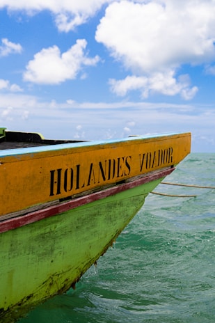 A wooden boat with the name 'Holandes Voador' painted on its yellow and green hull is floating in clear sea water under a bright blue sky with scattered white clouds.