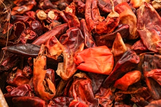 A close-up view of a pile of dried red chili peppers, showcasing a variety of shapes and textures. The peppers range in color from dark maroon to vibrant red and orange, with some peppers appearing wrinkled and dried out. Their surfaces have a glossiness that highlights their natural curves and edges.