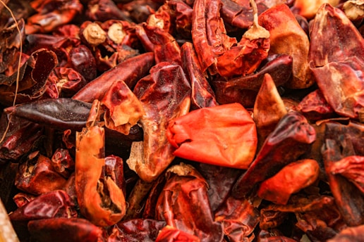 A close-up view of a pile of dried red chili peppers, showcasing a variety of shapes and textures. The peppers range in color from dark maroon to vibrant red and orange, with some peppers appearing wrinkled and dried out. Their surfaces have a glossiness that highlights their natural curves and edges.