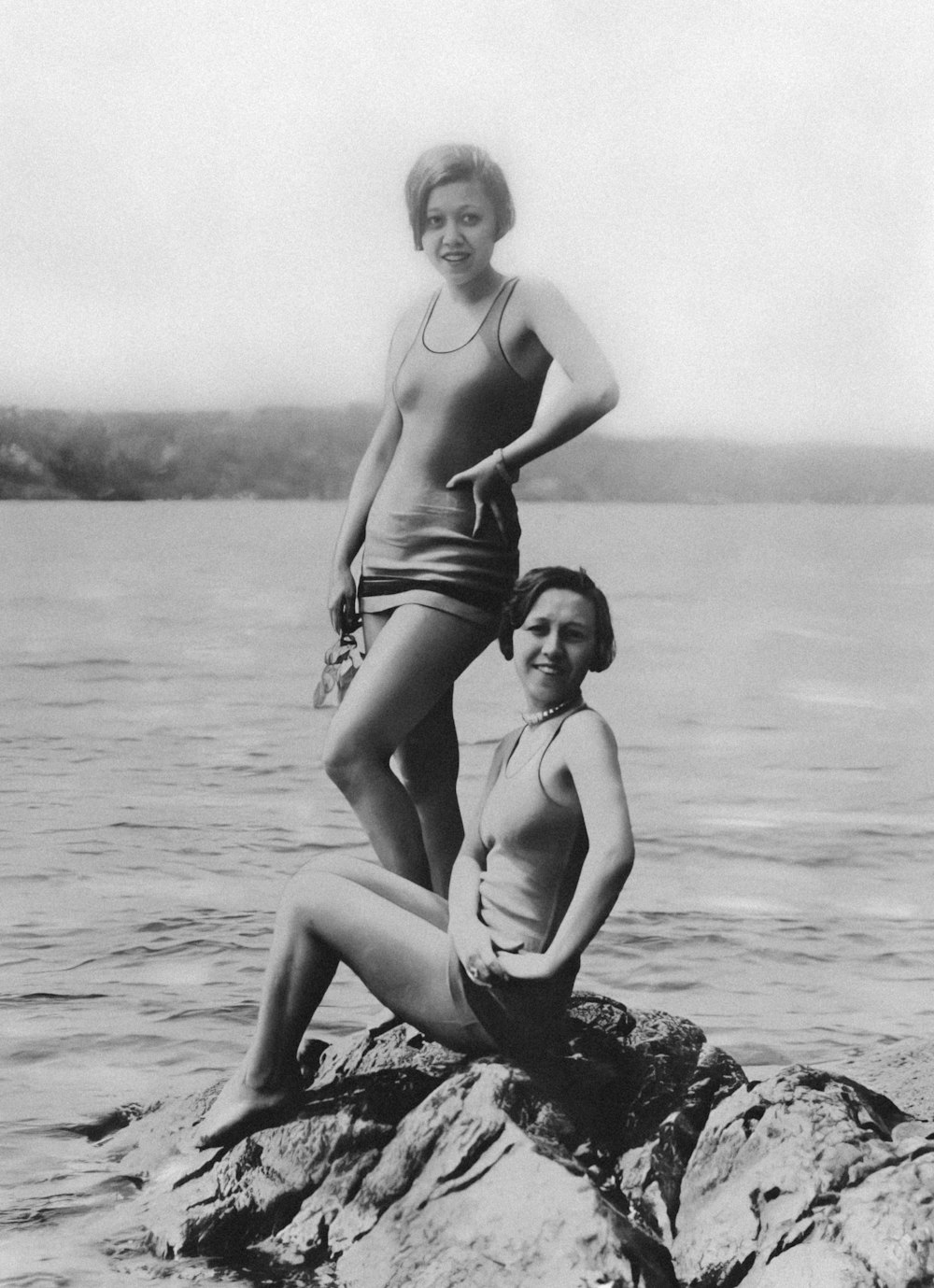 a couple of women standing on top of a rock next to a body of water