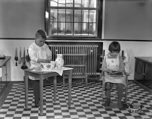 Two children are engaged in activities in a room with a checkered floor. A boy stands at a small table, working with a large, patterned bowl and a jug. A girl sits in a chair beside him, focusing on her task. The room features a large window, a radiator, and kitchen utensils hanging on the wall.