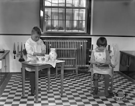 Two children are engaged in activities in a room with a checkered floor. A boy stands at a small table, working with a large, patterned bowl and a jug. A girl sits in a chair beside him, focusing on her task. The room features a large window, a radiator, and kitchen utensils hanging on the wall.