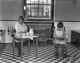 Two children are engaged in activities in a room with a checkered floor. A boy stands at a small table, working with a large, patterned bowl and a jug. A girl sits in a chair beside him, focusing on her task. The room features a large window, a radiator, and kitchen utensils hanging on the wall.