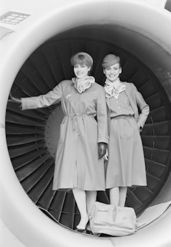 Two individuals are standing inside an aircraft engine nacelle. They are dressed in matching uniforms that consist of coats, scarves, and gloves, with one wearing a hat. A bag is placed on the ground beside them. The setting is monochromatic, focusing on the design of the engine.